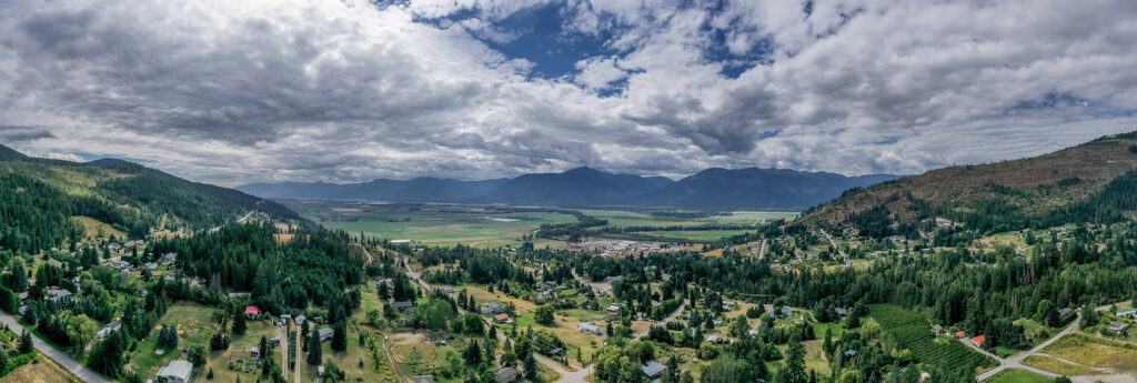 A panoramic photograph taken in Creston, BC, showcasing a sweeping view of a valley with residential and rural areas interspersed among patches of forest, surrounded by mountains and under a cloudy sky